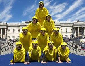 Ten performers, wearing yellow helmets, goggles & macs, kneel on each other's backs to create a pyramid