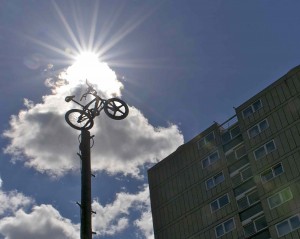 bmx bike leaps from the top of an old telegraph pole, the sun bursts from from a cloud behind against a blue sky. A tenement block sits in the corner of the image