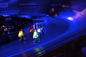 5 cyclists in green, red, yellow, white & blue helmets & lit up capes, with lights on their bikes & spokes cycle around a track in an arrow formation