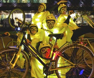 Five performers pose behind a bike with 2 bikes framing either side. Performers are dressed in yellow capes, helmets & goggles with lights underneath the campes on helmets & on the bikes