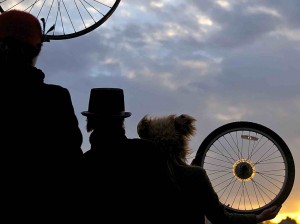 Three silhouetted figures gaze towards the sunset. Two hold bike wheels and each wears a different hat: a cloche hat with feathers, a top hat, and a furry bear's hat