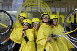Five performers wearing yellow macs, helmets & goggles pose in between two bikes raised on their back wheels
